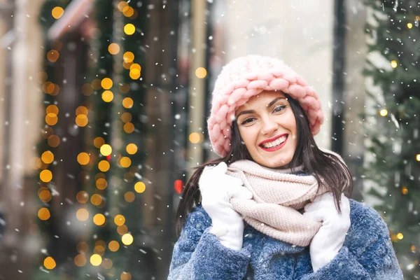 在降雪期间 快乐的黑发女人头戴针织浅粉色的帽子和围巾 走在装饰着花环的城市 案文的篇幅 — 图库照片