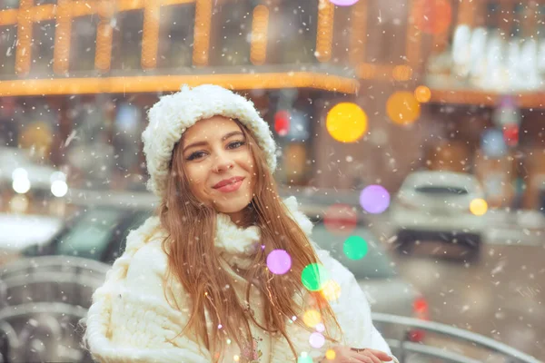 かわいい若い女性は白い帽子とコートを身に着けています雪の中で街の装飾されたガーランドを歩く — ストック写真