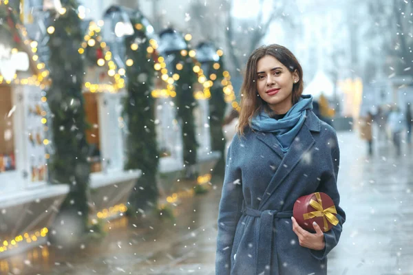 Güzel esmer kadın, kar yağarken Noel panayırının yanında hediye kutusu tutuyor. Metin için boşluk