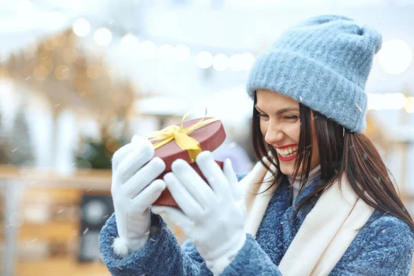 Kışlık ceketli, heyecanlı esmer kadın, kar yağışı sırasında Noel panayırında hediye kutusu tutuyor. Metin için boşluk