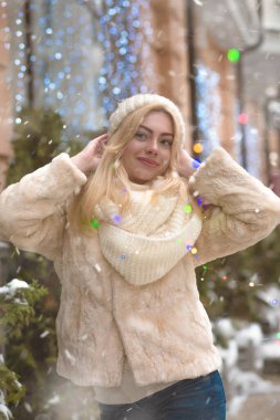 Güzel sarışın kadın kar yağışı sırasında renkli çelenklerin yanında yürüyor.
