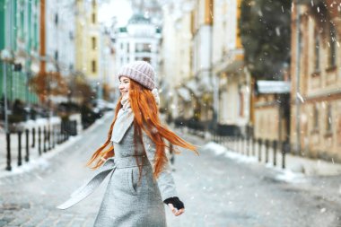 Kızıl saçlı mutlu kadın, Noel tatillerinde kar eşliğinde sokakta yürürken şık bir ceket giyiyor. Metin için boşluk