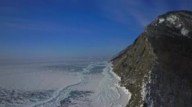 İnsansız hava aracı, kış ağaçlarıyla gri kayaların üzerinde dönüyor. Sonsuz buz kıyı şeridini ortaya çıkarıyor.