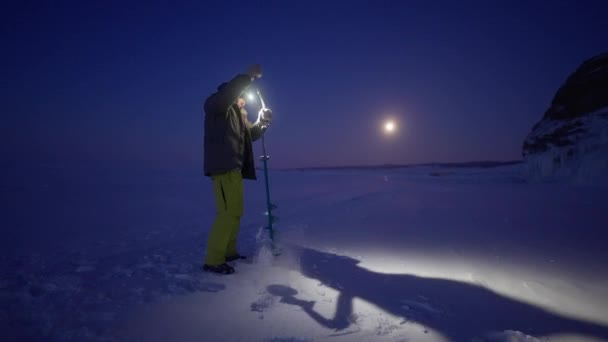Hombre taladro agujero con sinfín de mano en hielo congelado. La sombra gira en la luz de la cabeza — Vídeos de Stock