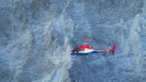 Helicóptero de rescate rojo y azul volando alto a lo largo de montañas rocosas bajo el sol. — Vídeos de Stock