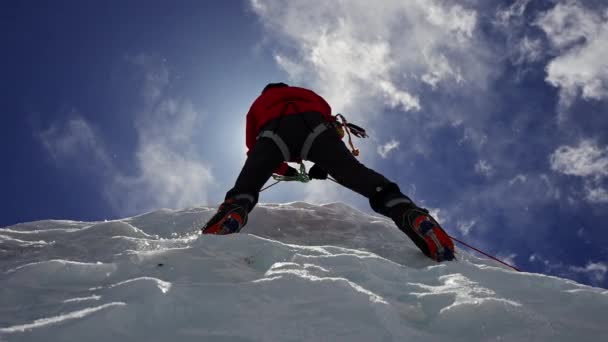 Kırmızı kramponla donatılmış dağcı mavi güneşli gökyüzünün altındaki buz duvarına tırman. — Stok video