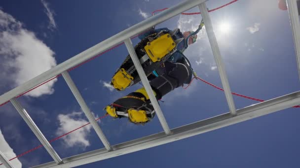 Sarı kramponla donatılmış dağcı mavi gökyüzünün altında asılı duran merdivenden geçiyor. — Stok video