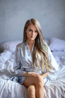 Attractive and sexy young blonde woman in silver nightgown sitting on the bed