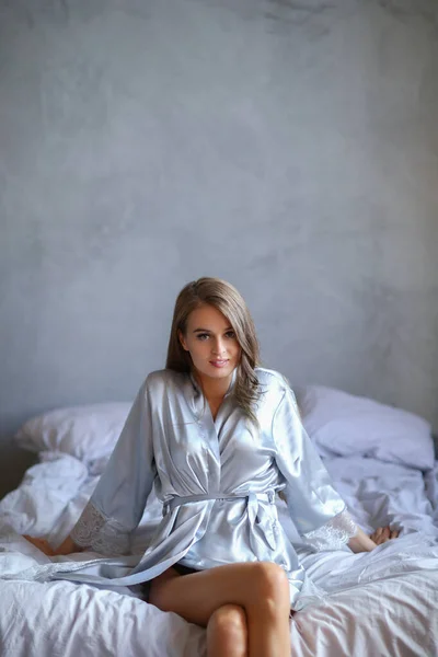 穿着银色睡衣坐在床上的年轻貌美性感的金发女人 — 图库照片