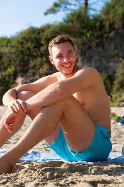 Oğlan ya da genç adam kumsalda güneşleniyor ve gülümsüyor.