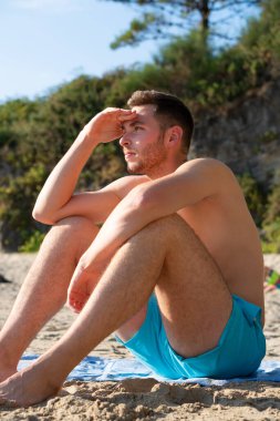 Genç ya da erkek, kumsalda oturmuş mavi mayo giymiş güneşleniyor.
