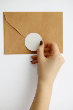 Manikürlü kadın eli beyaz arka planda kahverengi bir zarfa yuvarlak bir çıkartma yapıştırıyor. çerçeve kesme ile modelleme etiketleri
