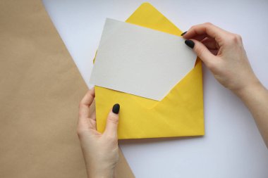 Beyaz bir çarşaf ve kahverengi beyaz bir zemin üzerinde sarı bir zarf tutan manikürlü kadın elleri. Kartpostal düzeni