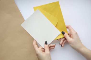 Beyaz çarşafı dik tutan manikürlü kadın elleri ve kahverengi beyaz arka planda sarı bir zarf. Kartpostal düzeni