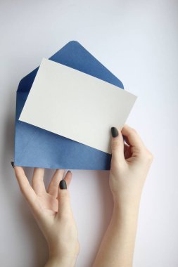 Manikürlü kadın elleri beyaz bir çarşaf ve mavi bir zarfı kahverengi ve beyaz arka planda yatay olarak tutuyor. Kartpostal düzeni