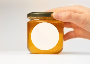 Bir kadının eli masanın üzerine bir kavanoz bal koyar. Üzerinde de entrikacı kesimi olan yuvarlak bir çıkartma. Paketleme tasarımı için boşluk ile