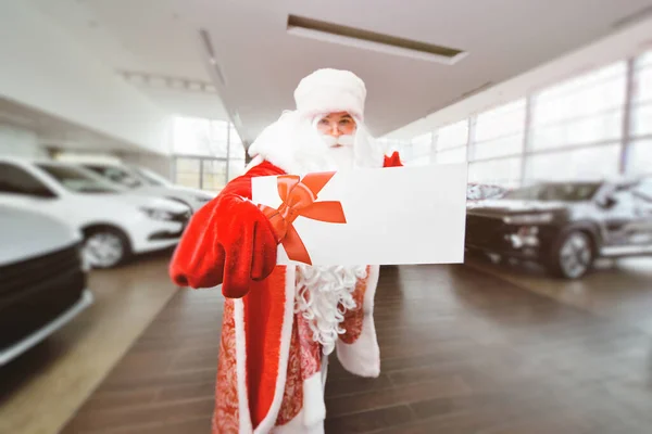 Papai Noel está segurando uma folha branca com um lugar para o texto no fundo de uma concessionária de carros. — Fotografia de Stock