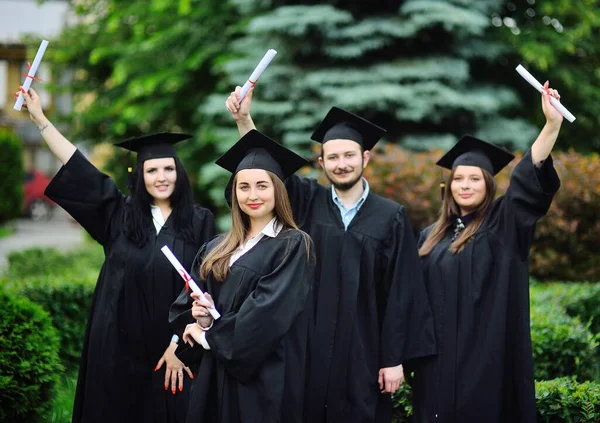 Jovem, uma estudante universitária de pós-graduação em um manto preto e um chapéu quadrado com um diploma em suas mãos, sorri contra o pano de fundo dos alunos colegas de classe. — Fotografia de Stock