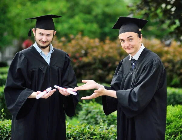Dos jóvenes graduados en túnicas y sombreros cuadrados están felices de recibir un diploma. —  Fotos de Stock