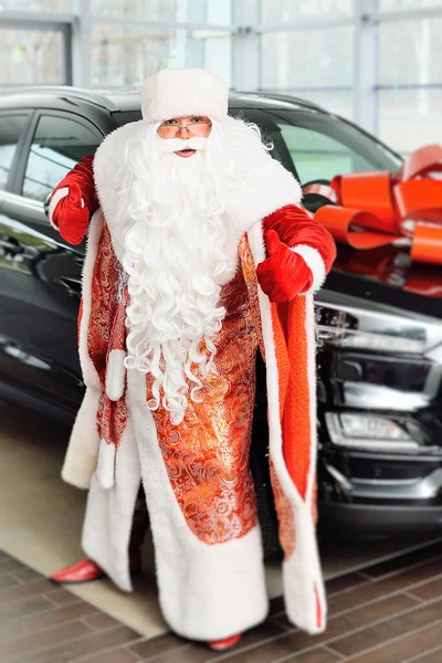 Papai Noel dá um polegar para cima contra o fundo de uma concessionária de carros e um novo carro preto com um arco vermelho. — Fotografia de Stock