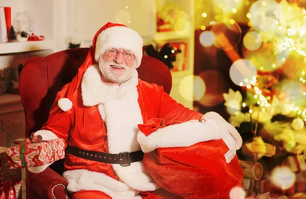Papai Noel com uma verdadeira barba branca em um terno vermelho e branco tradicional e óculos redondos está sorrindo sentado em uma cadeira e segurando um saco de presentes — Fotografia de Stock