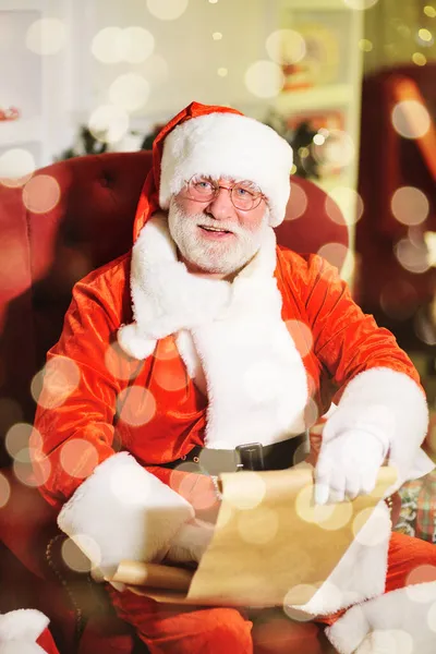 Papai Noel com uma verdadeira barba branca em um terno vermelho e branco tradicional e óculos redondos está sorrindo sentado em uma cadeira e segurando uma lista de desejos das crianças. — Fotografia de Stock