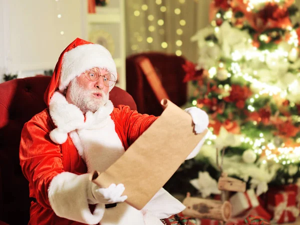 Papai Noel com uma verdadeira barba branca em um terno vermelho e branco tradicional e óculos redondos sentados em uma cadeira é surpreendido segurando em suas mãos uma longa lista com os desejos das crianças contra o — Fotografia de Stock