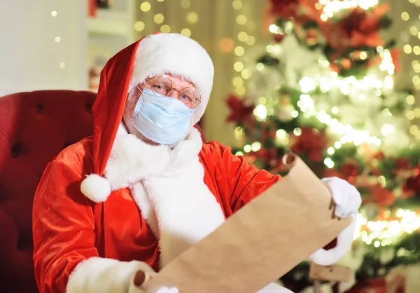 Papai Noel em uma máscara médica sentado em uma cadeira lendo a lista de desejos das crianças contra o fundo de uma árvore de Natal e decorações. — Fotografia de Stock