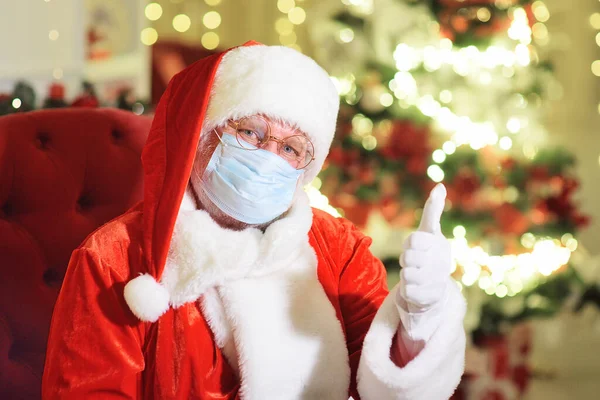 Papai Noel em uma máscara médica e luvas brancas sentadas em uma cadeira mostra um polegar para cima - um sinal de classe. — Fotografia de Stock