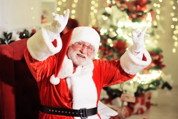 Alegre Papai Noel com uma verdadeira barba cinza em um traje tradicional e óculos redondos sorri e levanta as mãos contra o fundo de uma árvore de Natal. — Fotografia de Stock