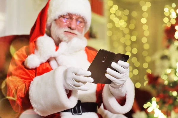 Papai Noel com uma barba cinza real em um traje tradicional está sentado em uma cadeira segurando um smartphone em suas mãos. Papai Noel no remoto congratula pessoas no Natal e Ano Novo por meio de social — Fotografia de Stock