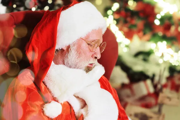 Retrato de Papai Noel com uma verdadeira barba cinza sentada em uma cadeira contra o fundo de uma árvore de Natal e luzes de Natal. — Fotografia de Stock