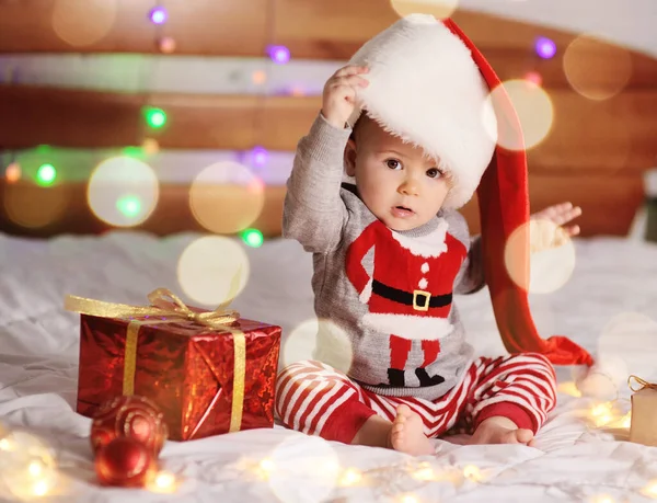 Bebé niño en un suéter de Navidad y un sombrero de Santa está sentado descalzo en una cama sobre un fondo de regalos y luces de Navidad —  Fotos de Stock