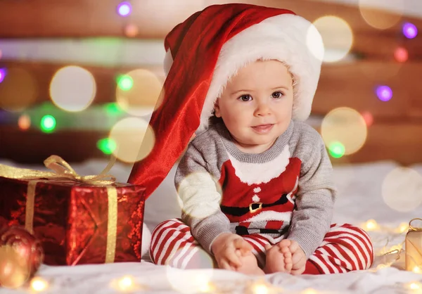 Bebé niño en un suéter de Navidad y un sombrero de Santa está sentado descalzo en una cama sobre un fondo de regalos y luces de Navidad —  Fotos de Stock