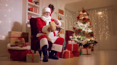 Noel Baba bir sandalyede oturmuş Noel ışıklarının arka planındaki çocuk dilekleri listesini okuyor.