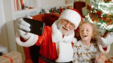 Gerçek gri sakallı Noel Baba küçük kıvırcık saçlı bir çocukla bir sandalyede oturuyor gülümsüyor ve akıllı telefondan selfie çekiyor.