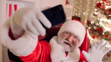 Noel Baba, elinde akıllı bir telefon tutuyor, gülümsüyor ve video iletişimiyle insanları Noel ve yeni yıl için tebrik ediyor.