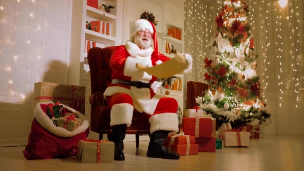 Santa con una barba blanca real en un traje rojo y blanco tradicional y gafas redondas está sonriendo sentado en una silla y sosteniendo una lista de deseos de los niños — Vídeo de stock