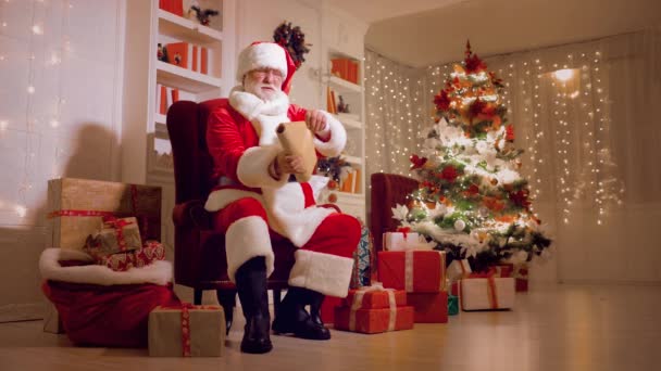 Санта Клаус сидит на стуле и читает список детских желаний на фоне рождественских огней — стоковое видео