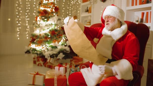 Santa Claus sedí na židli a čte si seznam dětských přání na pozadí vánočních světel — Stock video