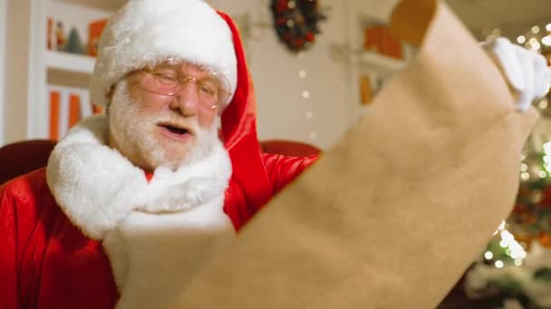 Santa Claus se skutečným bílým vousem v tradičním červenobílém obleku a kulatými brýlemi se usmívá na židli a drží seznam dětských přání — Stock video