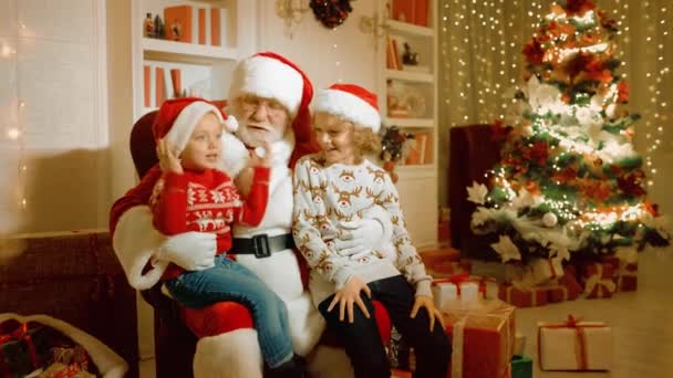 Crianças um menino e uma menina em camisolas de Natal estão segurando presentes em suas mãos e sentados em uma poltrona com Papai Noel — Vídeo de Stock