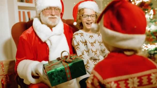 Niños un niño y una niña en suéteres de Navidad están sosteniendo regalos en sus manos y sentados en un sillón con Santa Claus — Vídeos de Stock