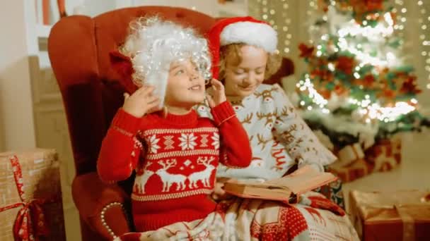 Ragazzo e una ragazza in maglioni di Natale con renne sono seduti su una sedia sullo sfondo di un albero di Natale, giocare, divertirsi, leggere un libro e mettere una parrucca di Babbo Natale sulla testa — Video Stock