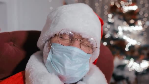 Papai Noel em um terno vermelho e branco tradicional e óculos redondos vai funcionar e coloca uma máscara médica descartável. — Vídeo de Stock