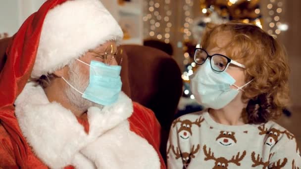 Санта Клаус и мальчик с вьющимися волосами в стерильных масках разговаривают на фоне рождественских огней — стоковое видео