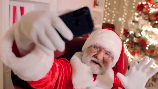 Santa Claus, sosteniendo un teléfono inteligente en sus manos, sonríe y felicita a la gente en Navidad y Año Nuevo a través de la comunicación por video — Vídeos de Stock