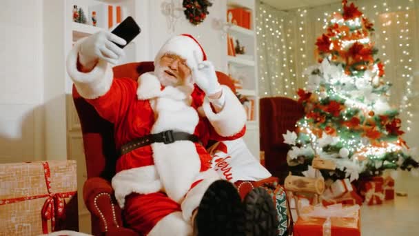 Санта-Клаус, держа в руках смартфон, улыбается и поздравляет людей с Рождеством и Новым годом с помощью видеосвязи — стоковое видео