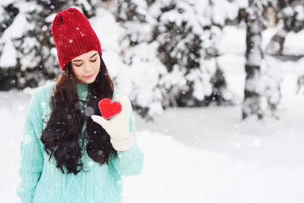 Молода красива дівчина в теплому синьому светрі і рукавицях на тлі засніжених дерев, що тримають червоне серце і посміхаються — стокове фото
