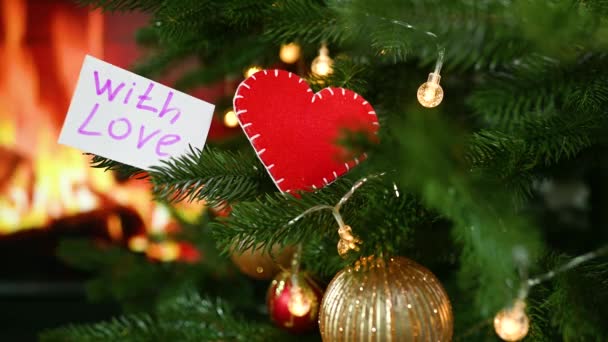 A red felt heart and a note with the text "With Love" on the background of a Christmas tree and a burning fireplace. — Vídeo de Stock